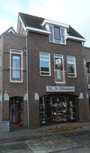 Een bakstenen gebouw van drie verdiepingen met een juwelierszaak op de begane grond en een "schilder" te koop bord in het raam van de middelste verdieping; de straat ervoor is nat, met wat gevallen bladeren verspreid over het trottoir.