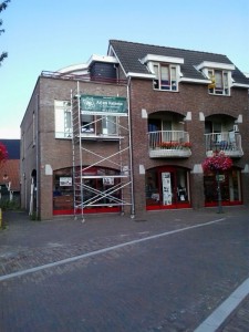 Een bakstenen gebouw met rode kozijnen en balkons toont een metalen steigerconstructie voor een nieuw project in aanbouw. Een groen "Ajan Rathma" bord hangt op de steiger, terwijl bloemen van de balkonhekken vallen.