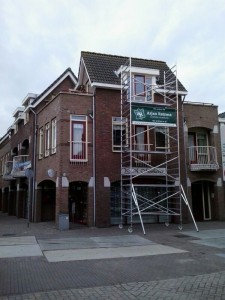 Een bakstenen gebouw met steigers voor een hoekingang markeert de start van een nieuw project. Op een groen bord op de steiger staat Arjan Ratema. Er zijn balkons en meerdere ramen op de bovenste verdiepingen.