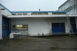 Een industrieel gebouw met witte en blauwe deuren wordt gedeeltelijk wit geschilderd. Steigers en een ladder staan opgesteld bij de muur en tonen de voortgang van het nieuwe schilderwerk op de rechterhelft van de gevel. Arjan Ratsma heeft het weer gefixt.