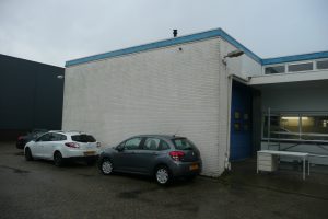 Twee auto's staan geparkeerd naast een eenvoudig wit bakstenen gebouw met een blauwe deur en kleine ramen. De grond is nat en er staat een buitenbalie bij de ingang. De lucht is bewolkt. Arjan Ratsma heeft het weer gefixt.