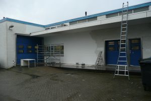 Een wit bakstenen gebouw met blauwe deuren wordt geschilderd. Ladders, steigers en schilderspullen staan buiten opgesteld en een deel van de muur is gedeeltelijk wit geschilderd. Een kleine tafel staat bij de deuren. Arjan Ratsma heeft het weer gefixt.