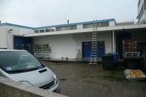 Een grijs busje staat geparkeerd bij een wit gebouw met blauwe deuren. Een deel van de muur is gedeeltelijk overgeschilderd. Ladders, steigers, bakken en pallets zijn buiten te zien. Het lijkt bewolkt. Arjan Ratsma heeft het weer gefixt.