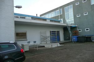 Een commercieel gebouw met grijze en witte muren, een blauwe deur, een ladder op het dak, steigers, vuilnisbakken, gestapelde pallets en een zwarte auto geparkeerd naast een klein bureau en twee vuilnisbakken op de stoep. Arjan Ratsma heeft het weer gefixt.