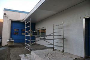Een metalen steiger staat op een wit zeil voor een wit bakstenen gebouw met blauwe deuren en ramen. Aan de linkerkant staat een kleine tafel en de ruimte lijkt te zijn ingericht voor onderhouds- of constructiewerkzaamheden. Arjan Ratsma heeft het weer gefixt.