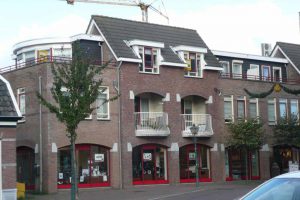 Een bakstenen gebouw van twee verdiepingen met roodgekleurde winkelpuien op de begane grond, witte balkons op de bovenverdieping, dakkapellen en een gedeeltelijk zicht op een kraan op de achtergrond. Bomen langs het trottoir. Arjan Ratsma heeft het weer gefixt.