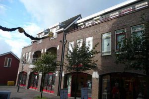 Een bakstenen gebouw van drie verdiepingen met boogramen en -deuren op de begane grond, balkons op de tweede verdieping en een schuin dak. Kleine bomen omzomen het trottoir aan de voorkant. De lucht is gedeeltelijk bewolkt. Arjan Ratsma heeft het weer gefixt.