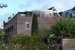 Een bakstenen gebouw van twee verdiepingen met rechthoekige ramen en een cirkelvormige structuur op het dak, gedeeltelijk verborgen door bomen en struiken op de voorgrond. Arjan Ratsma heeft het weer gefixt.