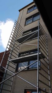 Een hoge metalen steigerstructuur staat tegen een licht bakstenen gebouw, reikt tot de derde verdieping, met platforms op twee verschillende niveaus. De lucht is gedeeltelijk bewolkt. Arjan Ratsma heeft het weer gefixt.