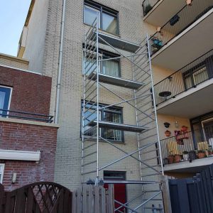 Voor een bakstenen woongebouw staat een metalen steigerconstructie die toegang biedt tot verschillende ramen op meerdere verdiepingen. Aangrenzende balkons tonen potplanten en verschillende buitenartikelen. Arjan Ratsma heeft het weer gefixt.