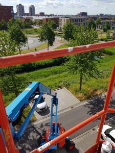 Zicht vanaf een verhoogde lift boven een straat, met de blauwe arm van de lift, een geparkeerde witte auto, bomen, gras en gebouwen op de achtergrond op een zonnige dag. De oranje veiligheidsreling is zichtbaar op de voorgrond. Arjan Ratsma heeft het weer gefixt.