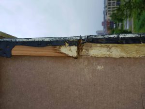 Close-up van een beschadigde dakrand met zichtbaar hout, afbladderende verf en tekenen van waterschade; gebouwen en groen zijn zichtbaar in de onscherpe achtergrond. Arjan Ratsma heeft het weer gefixt.