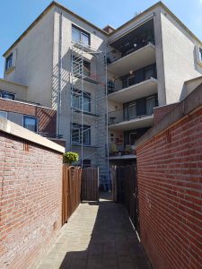 Een smal geplaveid steegje leidt naar een flatgebouw met balkons. Tegen de gevel van het gebouw staat een metalen steiger die van de grond tot het dak reikt. De lucht is helder en zonnig. Arjan Ratsma heeft het weer gefixt.