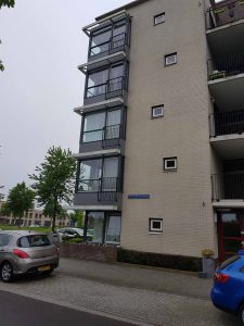 Een beige bakstenen flatgebouw met vier balkons is te zien bij een stoep op een bewolkte dag. Twee geparkeerde auto's en enkele bomen zijn zichtbaar langs de straat op de achtergrond. Arjan Ratsma heeft het weer gefixt.