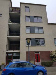 Een flatgebouw van drie verdiepingen met beige en bruine bakstenen muren, balkons aan de linkerkant en een rode deur naast een struik. Een blauwe auto staat geparkeerd op straat voor een lantaarnpaal. Arjan Ratsma heeft het weer gefixt.