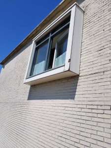 Een close-up van een modern, rechthoekig raam in een witte bakstenen muur. Eén ruit staat open en binnenin is een kort kanten gordijn zichtbaar. De lucht erboven is helder en blauw. Arjan Ratsma heeft het weer gefixt.