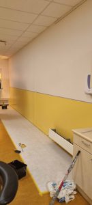 Een gang met een pas geverfde geel-witte muur, schilderstape langs de vloer, gereedschap, verfbenodigdheden en een gedeeltelijk afgedekte verwarming, wat duidt op renovatiewerkzaamheden. Arjan Ratsma heeft het weer gefixt.