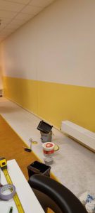 Een gedeeltelijk geverfde muur met een geel onderste gedeelte en een wit bovenste gedeelte. Schildergereedschap, een meetlint en een emmer staan op de vloer, die is afgedekt ter bescherming. Een radiator is aan de muur bevestigd. Arjan Ratsma heeft het weer gefixt.