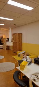 Een kinderdagverblijf of peuterspeelzaal met gele en witte muren, een houten kledingkast, een hangwieg, dozen met benodigdheden, een lage witte tafel met stoelen en ronde grijze vloerkleden op een geelbruine vloer. Arjan Ratsma heeft het weer gefixt.