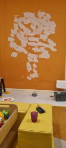 Een muur met een oranje achtergrond heeft witte plamuurvlekken in een ruwe boomvorm. Beneden op een tafel liggen schildergereedschap, een doosje, een spons, een beker en wat kinderspeelgoed op gele kussens. Arjan Ratsma heeft het weer gefixt.