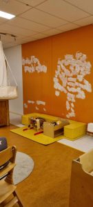 Een kinderspeelhoek met gele vloerkussens, houten speelgoed en een opbergdoos. De oranje muur erachter heeft meerdere witte vlekken, mogelijk van recente reparaties. Het plafond is wit met bovenlichten. Arjan Ratsma heeft het weer gefixt.