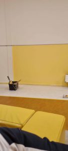 Een gedeeltelijk geverfde muur met een geel gedeelte in uitvoering. Een zwarte verfemmer met kwasten en een verfroller liggen op de grond. Gele kussens of banken zijn zichtbaar op de voorgrond. Arjan Ratsma heeft het weer gefixt.